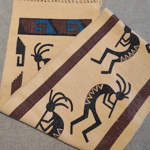 Puede incluir: Alfombra tejida beige con diseños geométricos y Kokopelli. La alfombra presenta un fondo beige con detalles en marrón y azul. Las figuras de Kokopelli son negras con detalles rojos. La alfombra tiene un borde con flecos.