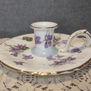 May include: A white porcelain candle holder with a circular base and a central socket. The piece features a painted pattern of purple violet flowers and green leaves, accented with a gold-trimmed scalloped edge and a curved handle with gold detailing.
