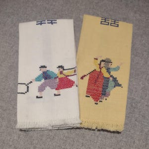Może przedstawiać: Dwa haftowane lniane ręczniki. Jeden jest w kolorze ecru z niebiesko-czarnym wzorem dwóch osób grających w grę. Drugi jest żółty z czerwono-niebieskim wzorem dwóch tańczących osób. Oba mają frędzle.