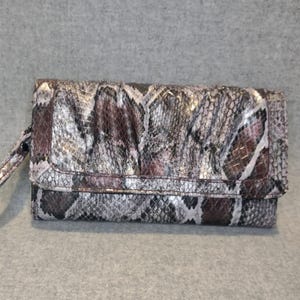 May include: A rectangular clutch wallet with a wrist strap. The wallet features a snakeskin pattern in shades of gray, brown, and silver. The flap closure is secured with a magnetic snap. The wallet is made of a textured material.