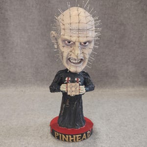 Może przedstawiać: Figurka typu bobblehead przedstawiająca Pinheada z bladą, pokrytą siatką głową pełną srebrnych szpilek, ubranego w czarną, teksturowaną szatę z czerwonymi akcentami. Postać trzyma małą kostkę logiczną i stoi na czerwonej, okrągłej podstawie z napisem.