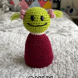 May include: Crochet pattern for a green and red doll with pink pigtails. The doll has a smiling face with black eyes. The pattern is for the doll only.
