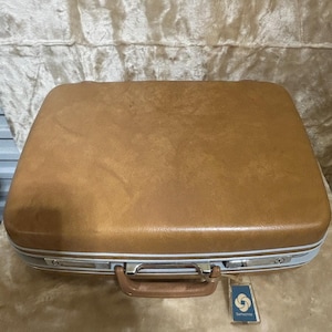 May include: A vintage Samsonite suitcase in a warm brown hue. The suitcase features a sturdy handle, silver-toned accents, and a light blue trim. A Samsonite tag hangs from the handle. The suitcase is rectangular and has a slightly textured surface.