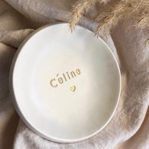 Peut inclure: Un vide-poches en céramique blanche avec le nom "Céline" en lettres dorées et un petit cœur doré. Le plat est rond et repose sur un tissu de couleur neutre, avec des plantes séchées en arrière-plan.