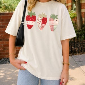 Puede incluir: Una camiseta de manga corta color blanco roto con un gráfico de tres fresas. Cada fresa está diseñada con un patrón de patchwork de telas rojas, blancas y verdes, incluyendo estampados florales, de cuadros vichy y de lunares, con hojas verdes.