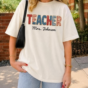 Puede incluir: Una camiseta de manga corta color blanco hueso con la palabra TEACHER en un diseño de letras de bloque con patrones rojos, blancos y azules. Debajo de la palabra, el texto Mrs. Johnson está impreso en una fuente cursiva negra al frente.
