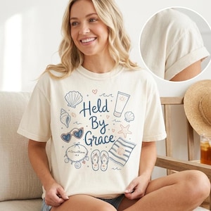 Puede incluir: Una camiseta de manga corta color crema con un estampado gráfico que incluye el texto "Held By Grace" rodeado de ilustraciones de conchas marinas, gafas de sol en forma de corazón, un protector solar, chanclas, una toalla y una pulsera.