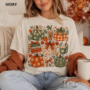 Puede incluir: Una camiseta color marfil con un estampado gráfico de varias calabazas, una pila de libros con las etiquetas cozy days, fall vibes, good books y pumpkin season, un vaso de café y una taza con un corazón, decorados con lazos, hojas y flores.
