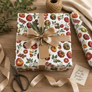 Puede incluir: Una caja de regalo rectangular envuelta en papel blanco con un patrón colorido de tomates rojos, naranjas y verdes en ramas. La caja está atada con una cinta de satén ancha y lisa de color marrón claro e incluye una etiqueta de regalo de papel kraft.