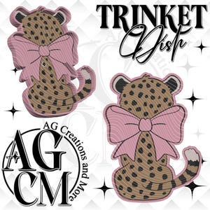 May include: A trinket dish featuring a cheetah design with a pink bow. The cheetah has brown spots and a striped tail. The words "TRINKET Dish" are above the cheetah. The logo "AG Creations and More" is also visible.