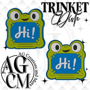 May include: Two identical frog-shaped trinket dishes featuring a bright green border with large eyes and a blue center panel. The word Hi! is written in white script in the center of each blue panel. The design includes small frog feet on the sides.