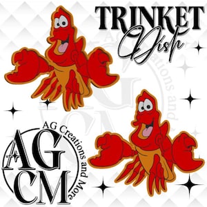 May include: A cartoon crab in red and orange, with the words "TRINKET Dish" in black script. The image also features the logo "AG Creations and More" in black and white. The crab is smiling with its tongue out.