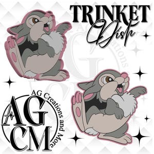 May include: A trinket dish featuring a cartoon rabbit character in shades of gray, pink, and white. The dish has a playful design with the words "Trinket Dish" in black script. The logo "AG Creations and More" is also visible.