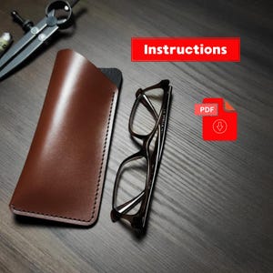 May include: A brown leather glasses case with visible stitching alongside a pair of dark-rimmed eyeglasses and a metal drafting compass. A red rectangular box contains the text Instructions and a red PDF download icon with a downward arrow.