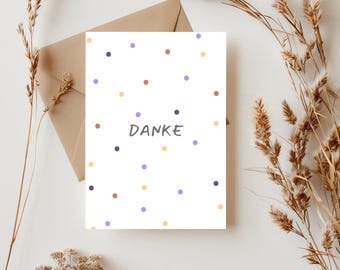 May include: A white card with a confetti pattern in shades of yellow, purple, blue, and red. The word "DANKE" is written in black in the center of the card.