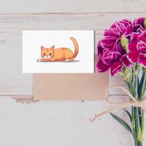 May include: A white card with a cartoon illustration of an orange tabby cat lying down. The cat has green eyes and a pink nose. The card is on a light brown surface.