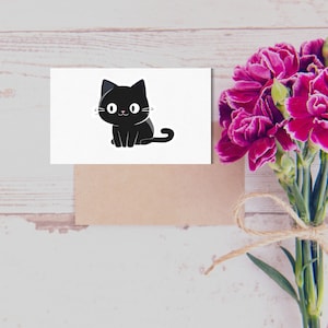May include: A white card with a black cartoon cat sitting on it. The cat has big eyes and a small smile. The card is sitting on a brown surface with a bouquet of pink flowers.