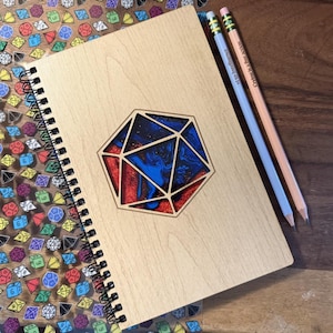 May include: A wooden spiral-bound notebook with a laser-cut geometric design filled with blue and red resin. Two pencils with text are placed on the right side of the notebook. The background features colorful dice.