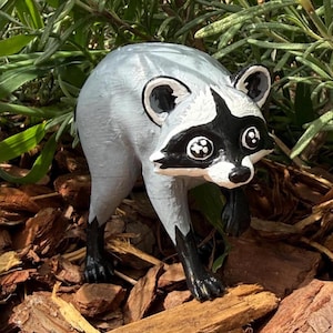 May include: A small raccoon figurine featuring a light gray body with black accents on the ears, eyes, and paws. The raccoon has a distinct black mask across its face, white fur around the eyes and muzzle, and large, round, dark eyes with white highlights.