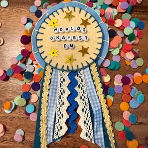 May include: A handmade felt award ribbon with the text "Worlds Okayest D.M." in white letter beads. The ribbon features blue and white gingham, lace, and gold star accents, surrounded by colorful felt confetti.