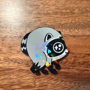 May include: A circular sticker or pin featuring a stylized raccoon illustration with a holographic finish. The design shows a gray raccoon body with black markings around the eyes, ears, and paws, and a striped tail, all rendered in a flat graphic style.