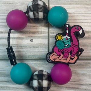 May include: A beaded bracelet featuring a flamingo charm holding a tropical drink. The bracelet includes magenta, teal, and black and white checkered beads. The bracelet is strung on a black cord.
