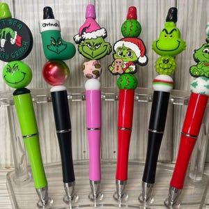 Op de afbeelding: Zes pennen met decoratieve toppers in Grinch-thema. De toppers bevatten een rond bordje met de tekst Don't Be A Salty Grinch, een groene kerstsok, een Grinch-hoofd met kerstmuts, een Grinch die een peperkoekmannetje vasthoudt en twee Grinch-figuren.