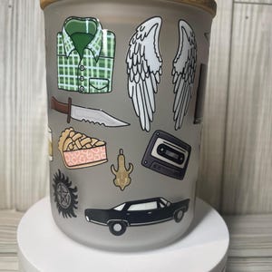 Puede incluir: Un vaso de vidrio esmerilado con tapa de bambú que presenta varias ilustraciones coloridas, incluyendo una camisa de cuadros verdes, alas de ángel blancas, un cuchillo plateado, un trozo de tarta, un casete negro, un colgante dorado y un coche negro.