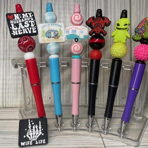 May include: A collection of decorative pens with various toppers, including a red heart-shaped sign that reads "N: MY HUSBAND'S LAST NERVE", a blue camper, and a green turtle. The pens are displayed in a clear stand.