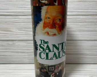 May include: A white tumbler with a collage of images from the movie "The Santa Claus". The images include Santa Claus, Tim Allen, and other characters from the film.