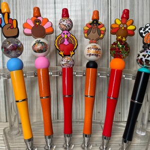 May include: Six pens featuring Thanksgiving-themed silicone toppers including turkeys, pumpkins, and a hand gesture. The pens have colorful barrels in yellow, orange, red, and black, accented with decorative beads in glittery, solid, and patterned finishes.