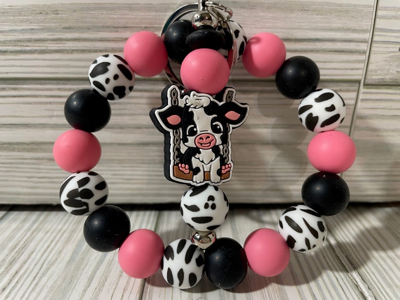 Puede incluir: Un llavero de pulsera con cuentas con un dije con tem&aacute;tica de vaca. La pulsera est&aacute; hecha de cuentas rosas, negras y con estampado de vaca. El dije de vaca es blanco y negro con una cara sonriente. Se adjunta un llavero plateado.