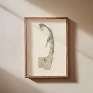 May include: Framed print of Cape Cod, Massachusetts, in a light brown wooden frame. The map is in shades of gray and beige, with the text "CAPE COD" and "MA" visible. The print is rectangular and hangs on a neutral-colored wall.