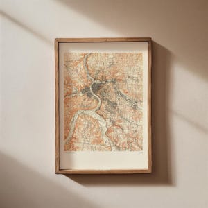 Puede incluir: Un mapa vintage enmarcado de Cincinnati, Ohio, con un marco de madera marrón y un fondo blanco. El mapa muestra las calles y los ríos de la ciudad en detalle, con un enfoque en el centro de la ciudad. El mapa está impreso en tonos de marrón, naranja y gris.
