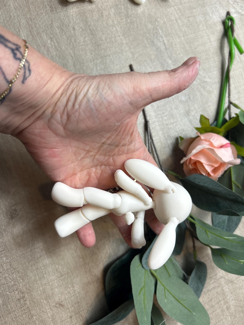 May include: A white articulated doll shaped like a rabbit with long ears. The doll is posed with its arms outstretched and its legs bent.