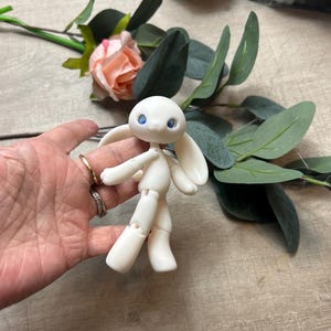 May include: A white articulated doll with blue eyes and bunny ears. The doll is posed with its arms outstretched and its legs bent.