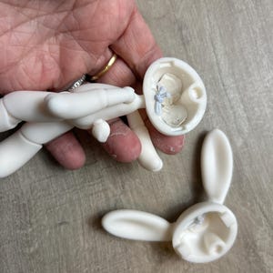 May include: White unfinished ceramic bunny rabbit figurine in pieces. The figurine is disassembled into three parts: the head, the body, and the two ears.