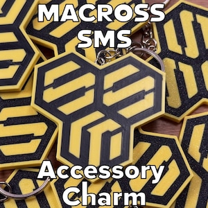 May include: Yellow and black accessory charms with the text "MACROSS SMS Accessory Charm". The charms have a geometric design and are attached to keychains. The charms are on a wooden surface.