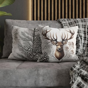 May include: Two decorative throw pillows on a gray sofa. One pillow features a black and white pine tree design. The other pillow displays a detailed illustration of a deer with large antlers, surrounded by bare branches and foliage.