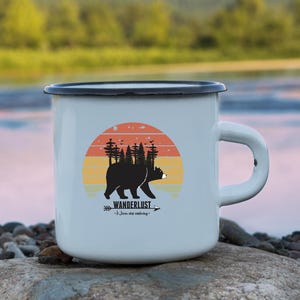 Può includere: Una tazza di smalto bianco con bordo e manico neri. La tazza presenta una grafica di un orso nero che cammina in una foresta con uno sfondo di tramonto. Il testo "Wanderlust" e "Just keep exploring" è stampato sulla tazza.