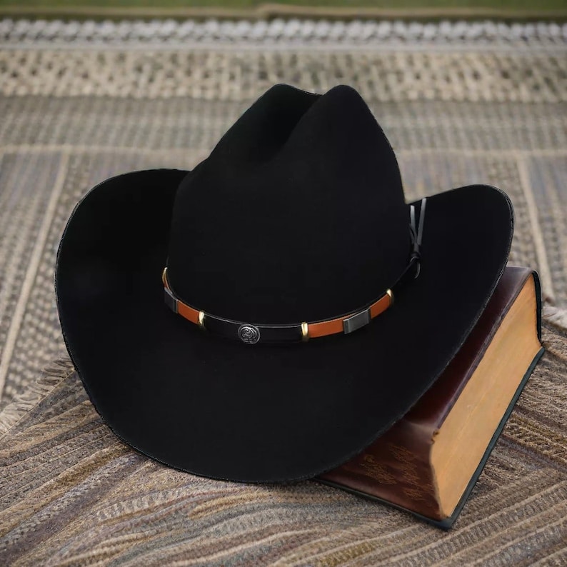 Pode incluir: Um chap&eacute;u de cowboy preto com uma faixa de couro marrom e detalhes em metal. O chap&eacute;u est&aacute; sobre um livro marrom.