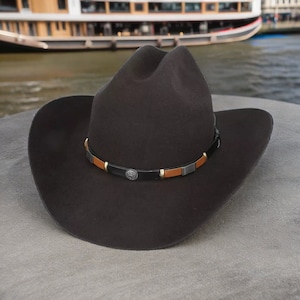 Pode incluir: Um chap&eacute;u de cowboy de feltro preto com uma faixa de couro marrom e detalhes prateados.