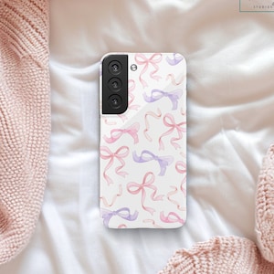 May include: A white phone case with a pattern of pink and purple watercolor bows. The case has a camera lens cutout and a black border.