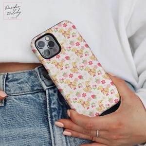 May include: A white phone case with a pattern of yellow ducklings and pink strawberries. The case is made of a hard plastic material and has a raised lip around the camera lens to protect it from scratches.