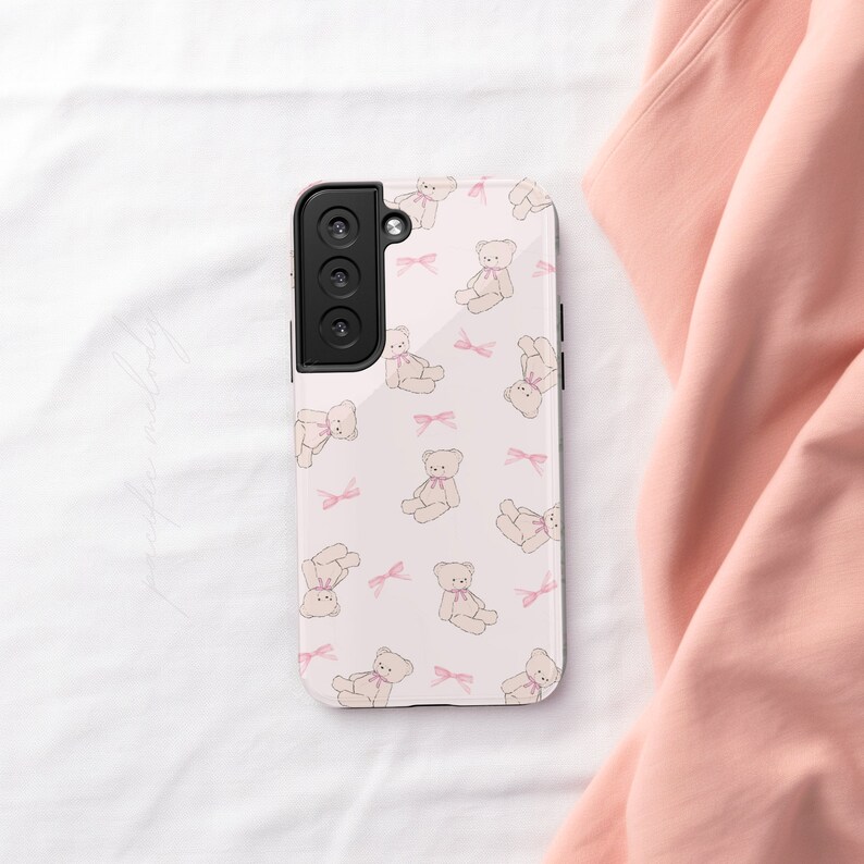 May include: A white phone case with a pink and white teddy bear pattern. The case has a black camera lens on the back.