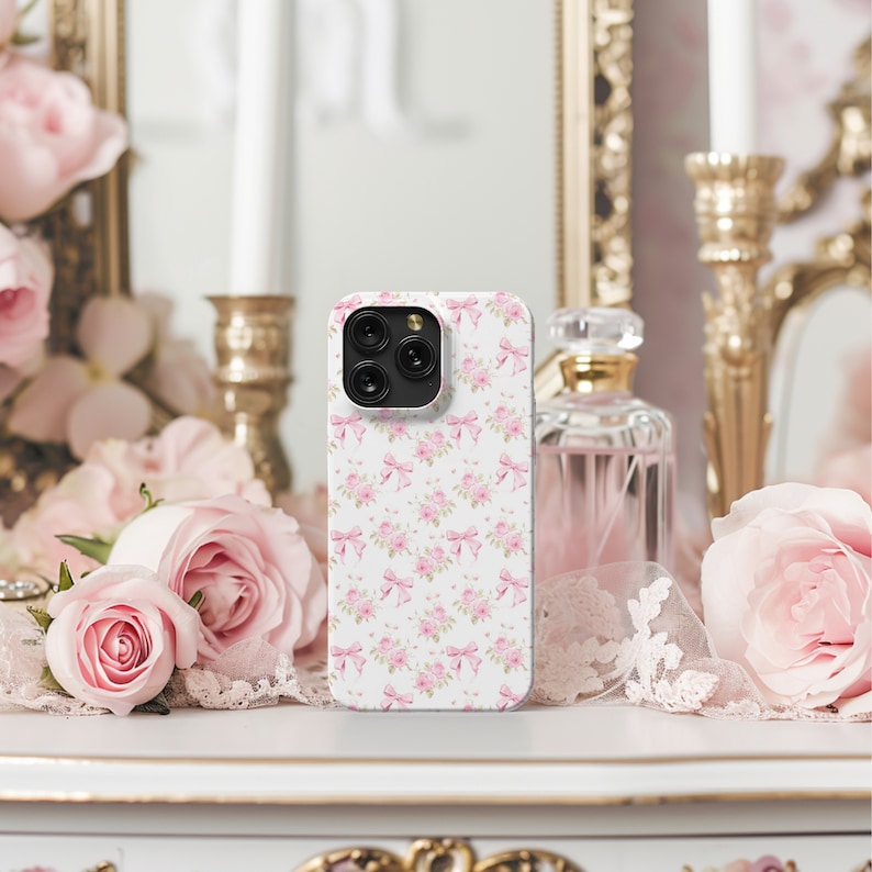 May include: A white iPhone case with a pink floral pattern featuring bows and roses. The case is on a white surface with pink flowers in the background.