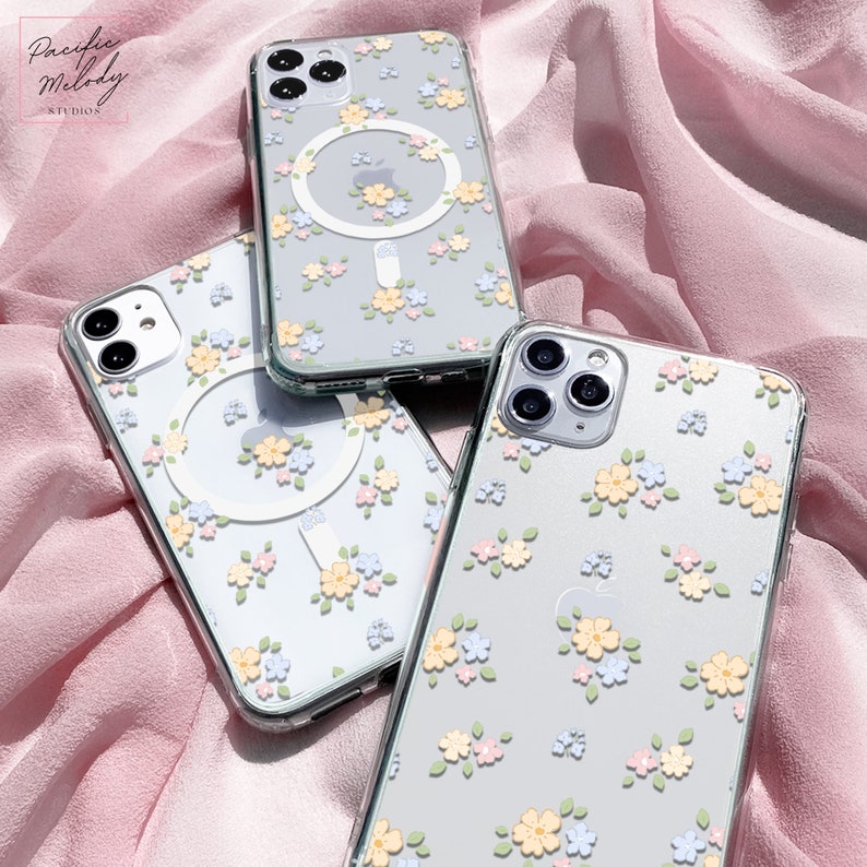 May include: Three clear phone cases with a floral pattern and a ring holder. The cases are on a pink fabric background.