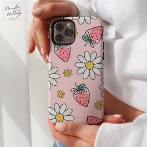 May include: A pink phone case with a white daisy and strawberry pattern. The case is designed for an iPhone with a camera bump.