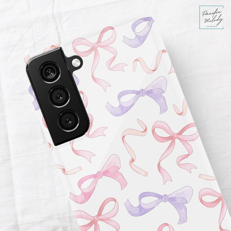 May include: A white phone case with a pattern of pink and purple watercolor bows. The case has a black camera lens cutout.