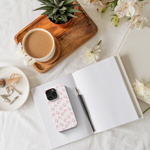 May include: A white iPhone case with a pink and gold bow pattern. The case is lying on a white surface with a notebook and a cup of coffee.
